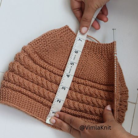 Hands measuring a brown hand-knitted cable beanie pattern with a measuring tape to check the correct hat length.