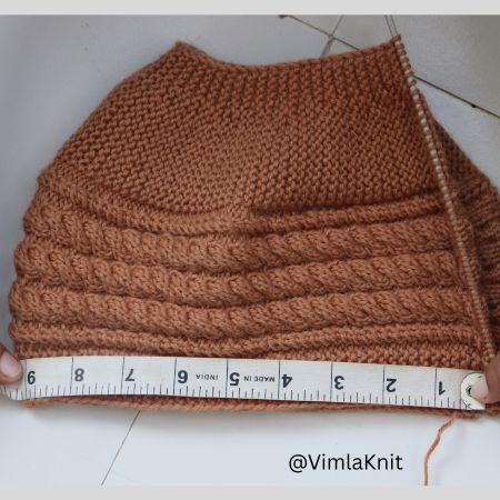 Hands measuring a brown hand-knitted cable beanie pattern on straight needles with a tape measure.