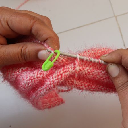 Close-up of hands knitting a pink twisted rib beanie, showing the twisted rib stitch pattern in progress with metal knitting needles, fuzzy yarn texture, and a green stitch marker, illustrating a handmade knit hat technique for a twisted rib beanie knitting pattern.
