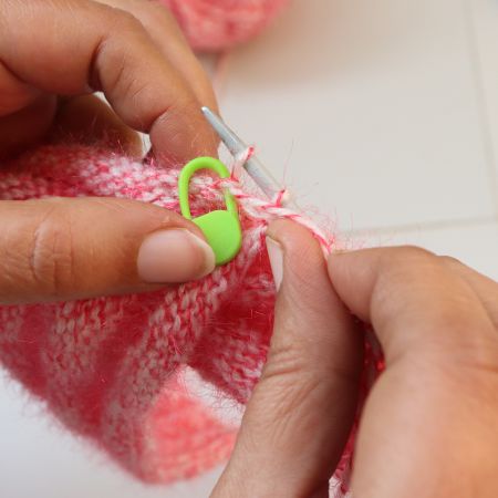 Close-up of hands knitting a pink twisted rib beanie, focusing on yarn tensioning and twisted rib stitch technique with metal knitting needles and a green stitch marker, highlighting handmade knit hat construction and twisted rib beanie knitting pattern details.