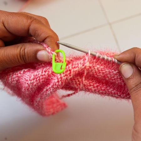 Close-up of hands knitting a pink twisted rib beanie pattern with metal knitting needles and a bright green stitch marker, showing detailed ribbed texture, yarn fibers, and in-progress handmade knit hat construction.