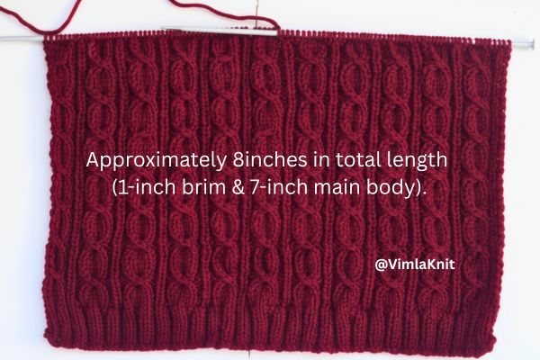 Maroon cable knit beanie body on knitting needles showing vertical twisted cable pattern, ribbed brim, and labeled measurements of approximately 8 inches total length, illustrating hat size and construction by Vimla Knit.