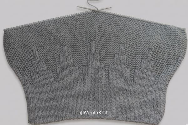 An in-progress view of a grey hand-knitted beanie on circular needles, resting against a seamless neutral background. The fabric features a defined ribbed brim and an architectural "skyline" rib pattern transitioning into garter stitch.