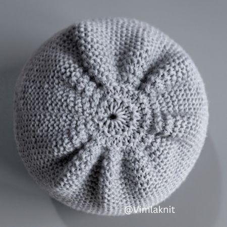 A high-resolution, overhead photograph showing the flawlessly decrease crown of a light grey knitted unisex beanie. The symmetrical knit decreases form a star-shaped pattern that perfectly closes the top of the hat, set against a clean, neutral grey studio background. The stitch definition is sharp, showcasing the expert finishing technique.
