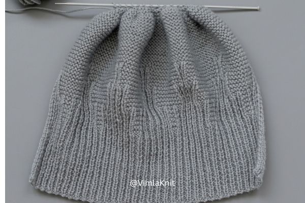 Close-up showing the crown shaping and decrease section of a grey knitted beanie. The live stitches are held on a knitting needle, with the yarn shown just before pulling through for a clean finish, against a grey background with the VimlaKnit watermark.