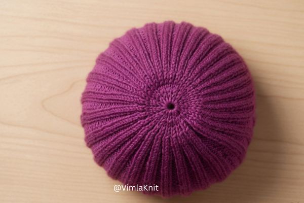 Top-down view of a purple ribbed knit beanie hat crown showing neat star-shaped decreases and a flat finish on a light wooden background.