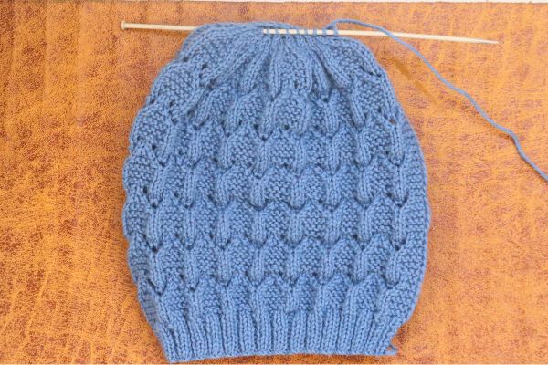 Blue lace knit beanie hat on knitting needles showing full diamond lace pattern and ribbed brim – Easy Knit Beanie Hat Pattern by Vimlaknit.