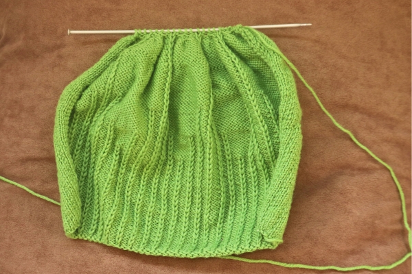Green knit beanie in progress on straight needles, showing rib and garter stitch pattern before crown shaping.