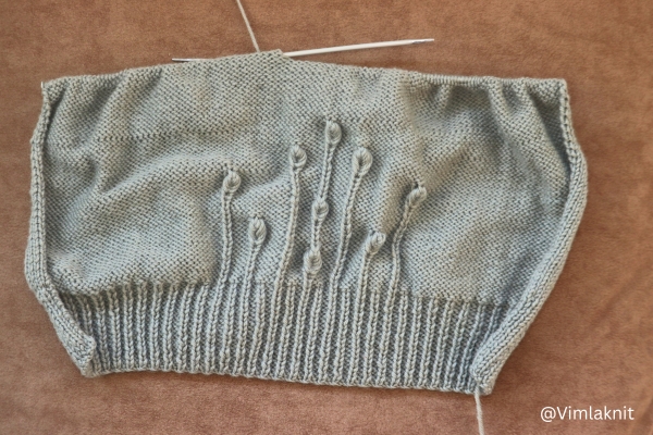 Light sage green knit beanie in progress on needles, showing ribbed brim, rib stems and raised leaf stitch pattern, leaf beanie knitting pattern.